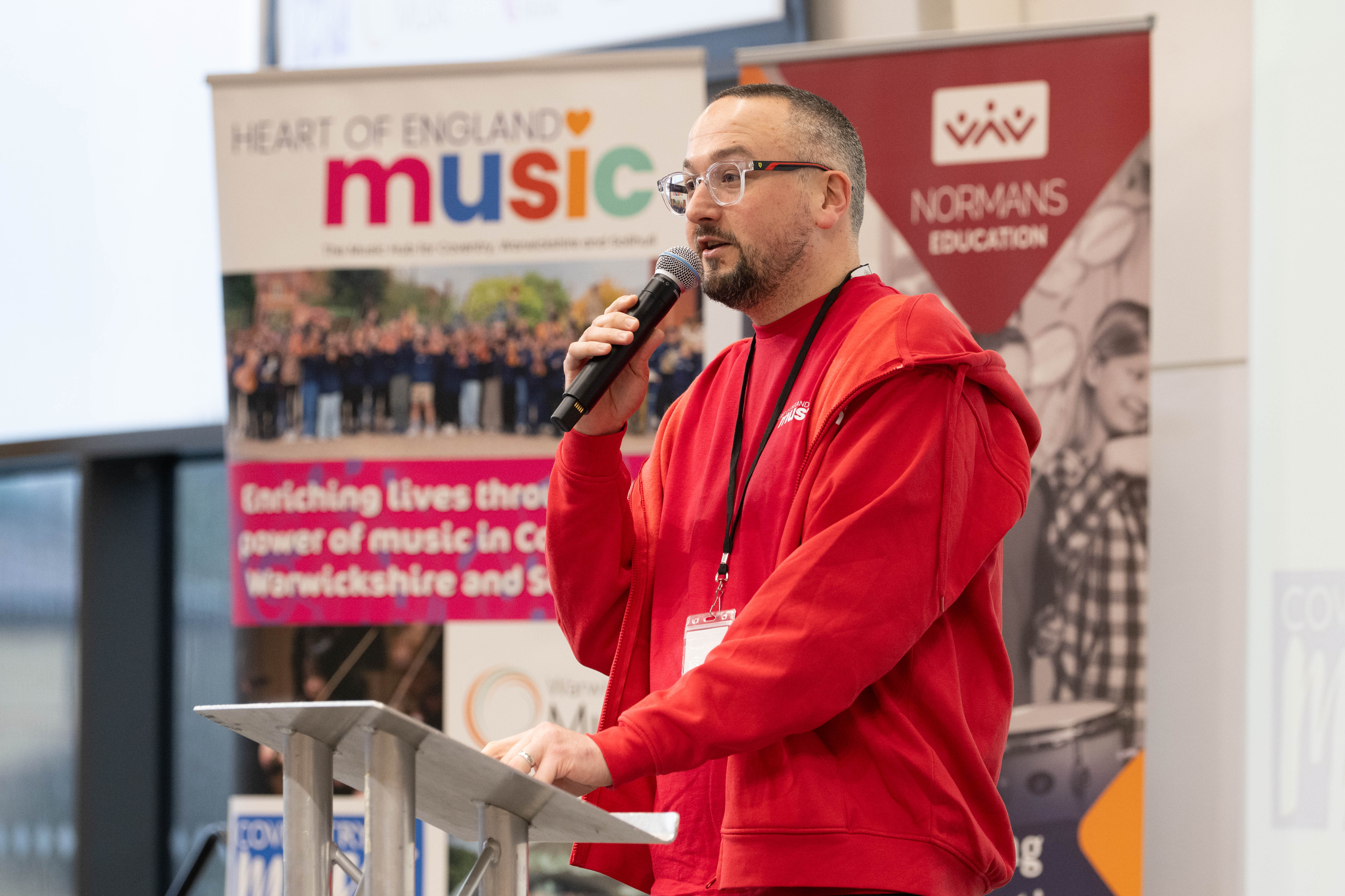 Mark Steele - Coventry Music Lead sanding on a stage with a microphone wearing a bright red hooded jacket presenting at a conference