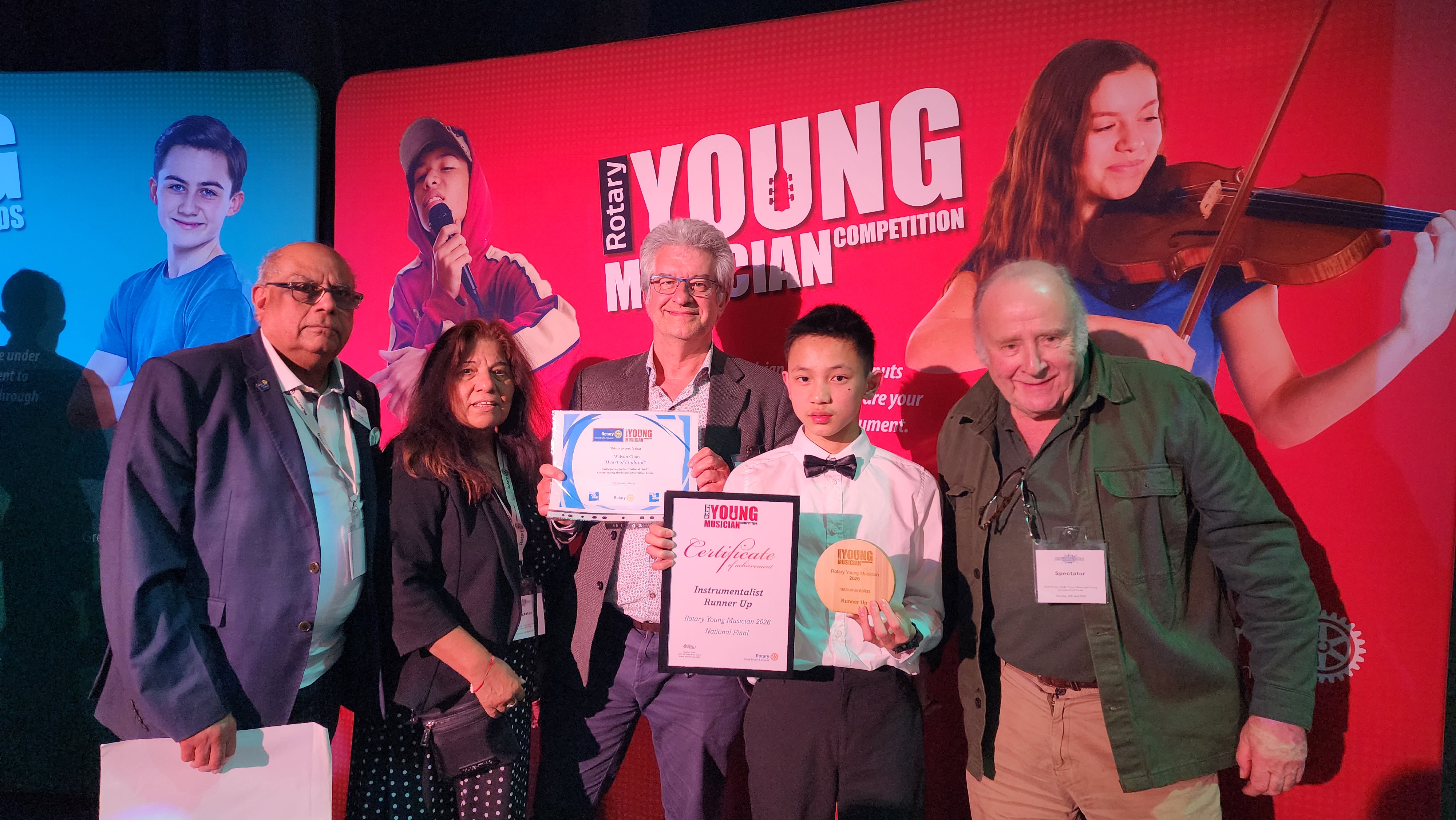 young musician in a bow tie proudly showing off his runner up certificate on a stage surrounded by his teacher and other event organisers