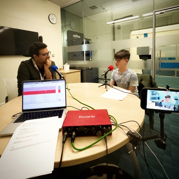 a young person and a teacher sat behind a desk with microphones recording a podcast
