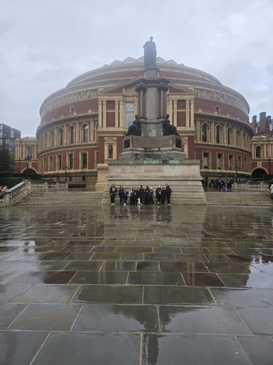 Picture of the Royal Albert Hall from a distance with a reflection on the wet floor and a group of people standing in front of the building