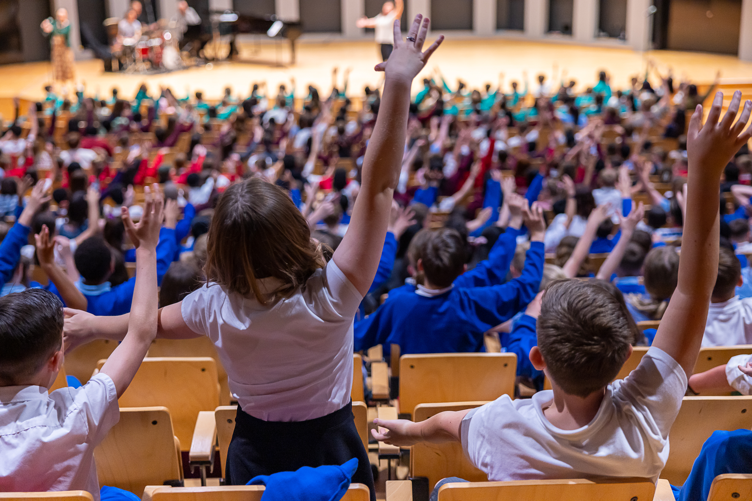 children singing in a theatre dressed in school unforms with their arms in the air