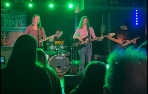 children playing drums, guitar and singing on a green lit stage with an audience looking on