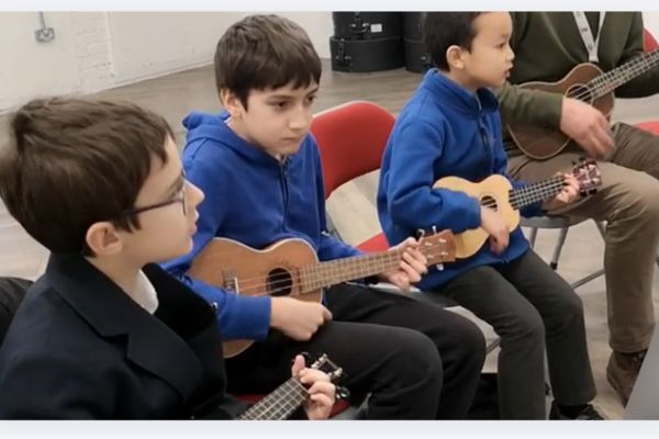 3 young children sitting on chairs playing ukuleles with their teacher