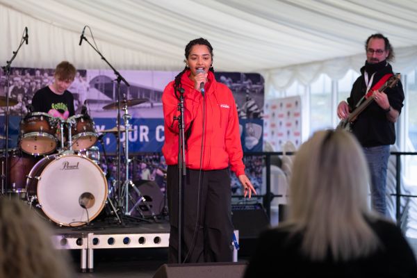 young adult singing in a red jacket, man playing electric guitar and boy playing drum kit on a stage